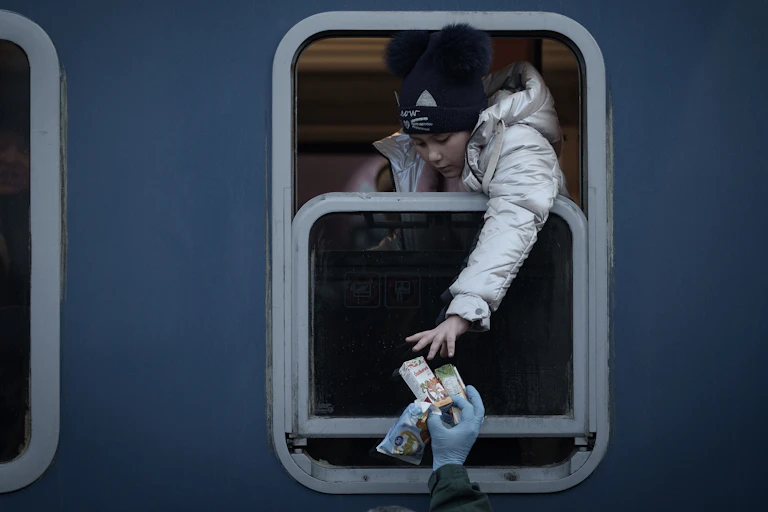 Person on train in silver jacket reaches out of window to take food from another person