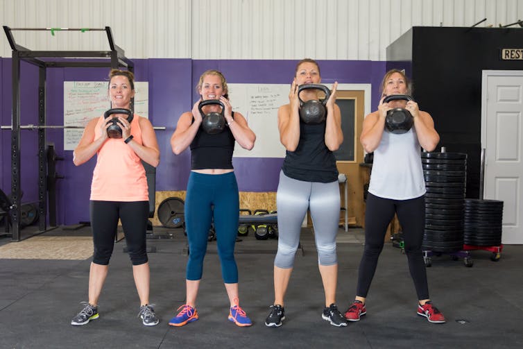 Four women in exercise clothes in a gym, holding kettle bells