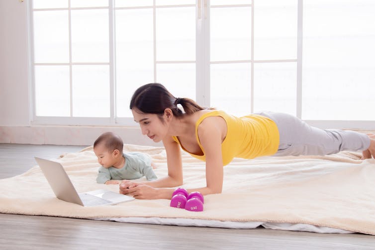 Woman doing a plank in front of an open laptop with her baby beside her and a pair of small dumbbells.
