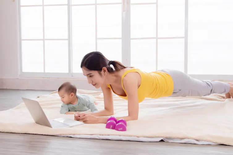 Woman doing a plank in front of an open laptop with her baby beside her and a pair of small dumbbells.
