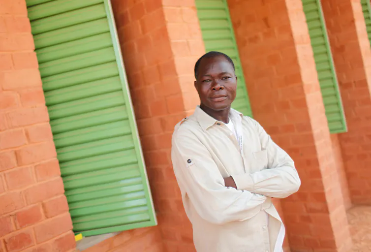 A man in a white shirt stands in front of an orange brick building with green shutters
