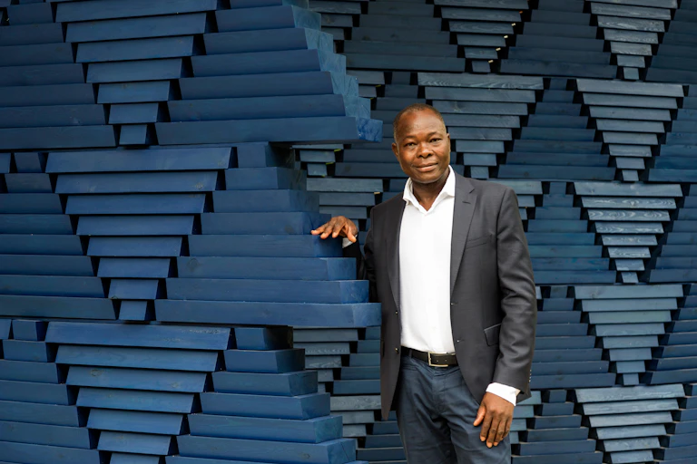 A man in a black jacket and white shirt stands in front of a dark blue sculptural façade.