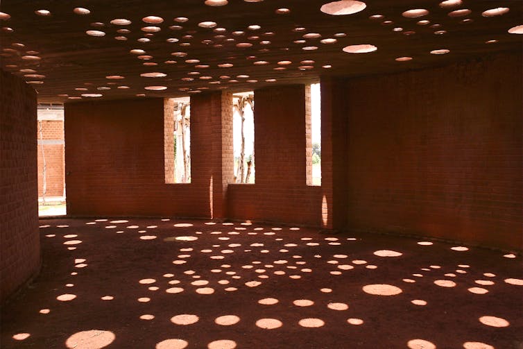An interior shot of a clay brick building with circular holes in the ceiling, through which light filters down onto the floor.