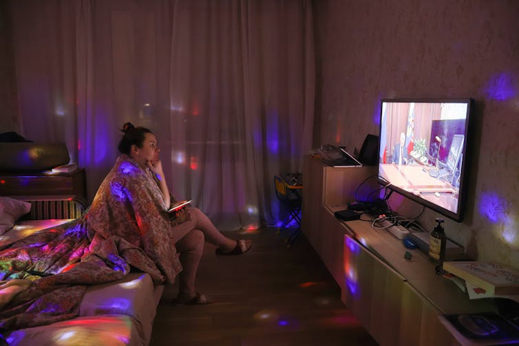 A woman sits in a dark room watching Russian President Vladimir Putin speak from an official looking desk