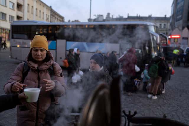 A woman reaches out her hand for a cup of soup, steam is coming from a big pot, a young boy stands next to her as people board coach busses in the background.