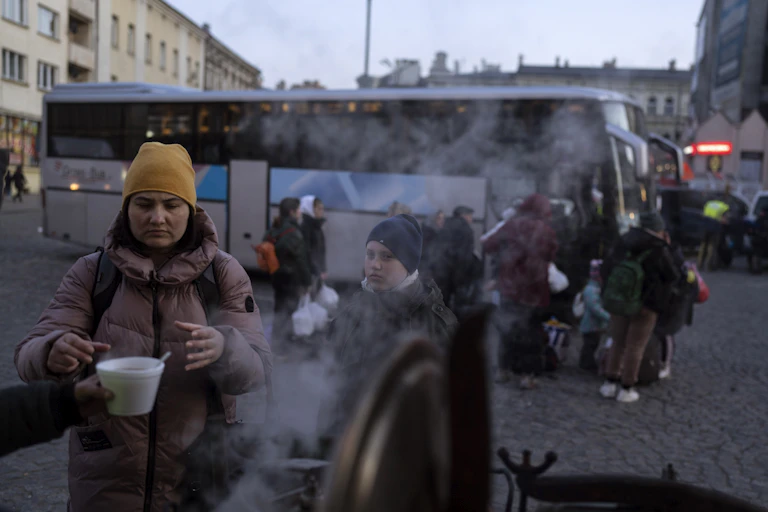 A woman reaches out her hand for a cup of soup, steam is coming from a big pot, a young boy stands next to her as people board coach busses in the background.