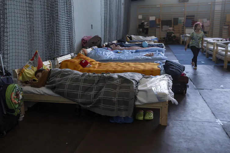 People sleep on beds in a row on the floor of a stadium.