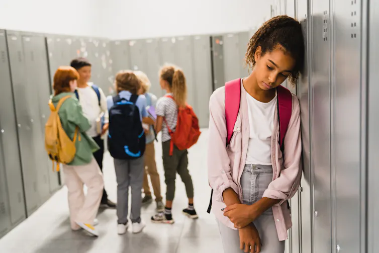 A group of school children talk in a circle while another student stands alone, leaning on a school locker.