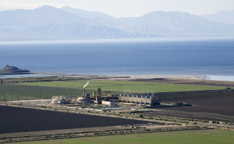 A power plant surrounded by fields with a large lake behind it and mountains in the distance.