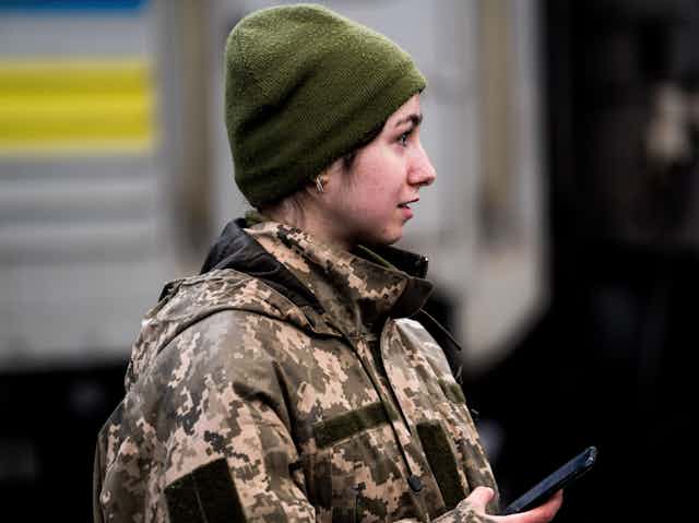 A young woman in a camouflage military uniform, holding a cellphone and wearing a green hat. 