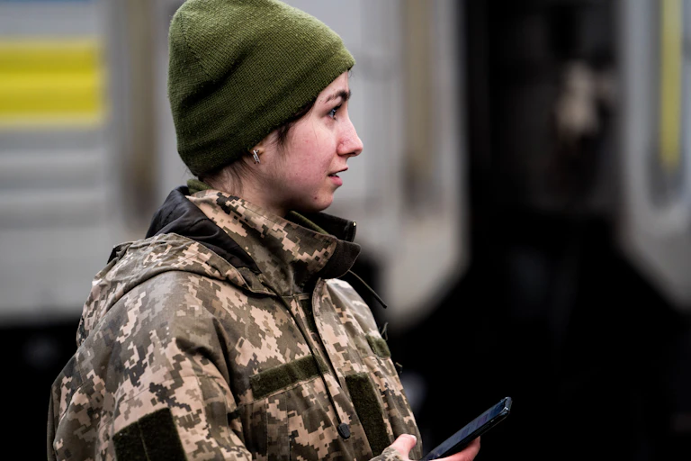 A young woman in a camouflage military uniform, holding a cellphone and wearing a green hat.