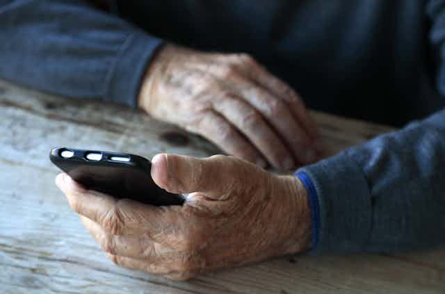 A person's arms in dark blue sweater sleeves are resting on a surface, with one hand holding a black smartphone.
