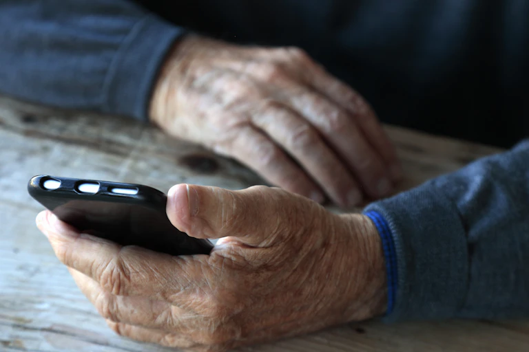 A person's arms in dark blue sweater sleeves are resting on a surface, with one hand holding a black smartphone.