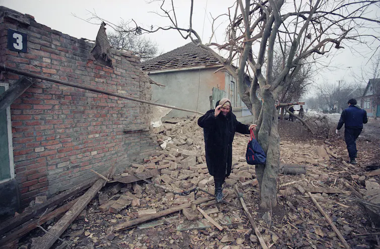 A grey-haired woman cries and holds onto a tree amid rubble.