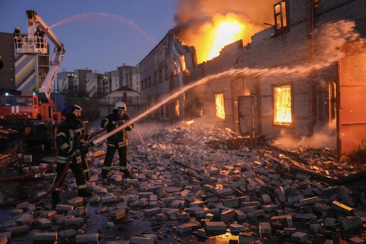 Firefighters hold a hose and spray water at a building burning ferociously with rubble strewn along the street