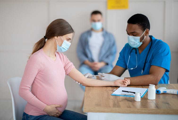 A pregnant woman sitting a table across from a health-care worker in scrubs and a face mask who is preparing to give her a shot.