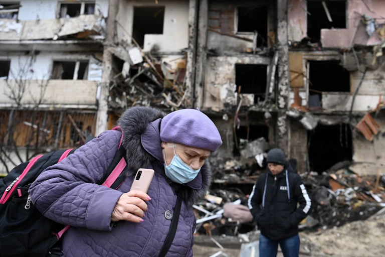 Une vieille dame masquée traverse une rue en ruine.