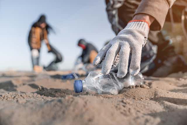 A gloved hand reaches down to pick a plastic bottle up off the sand.
