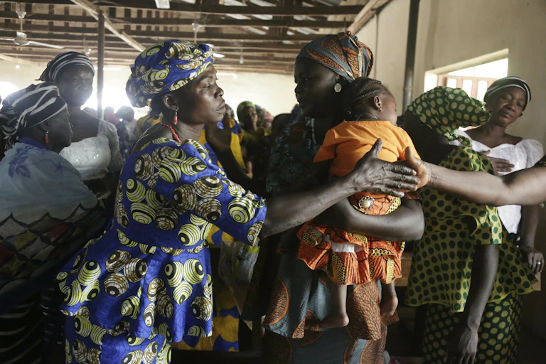 Women in colorful dresses shake hands in a crowd indoors.