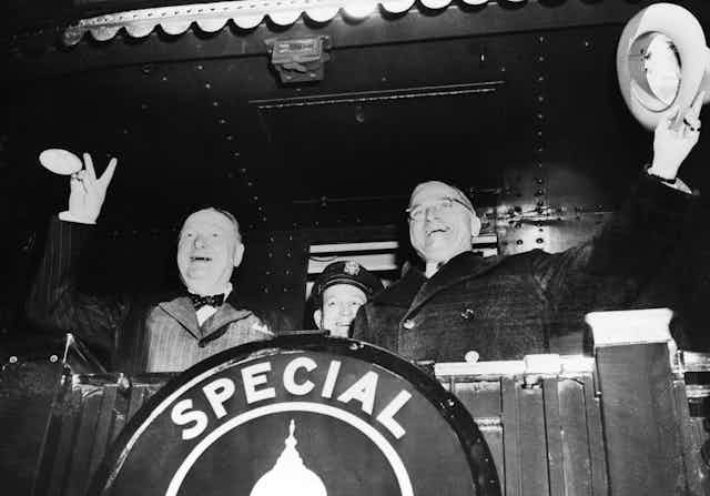 Two men on the back of a train, waving and smiling as it pulls out of the station.