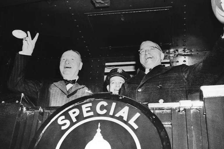 Two men on the back of a train, waving and smiling as it pulls out of the station.
