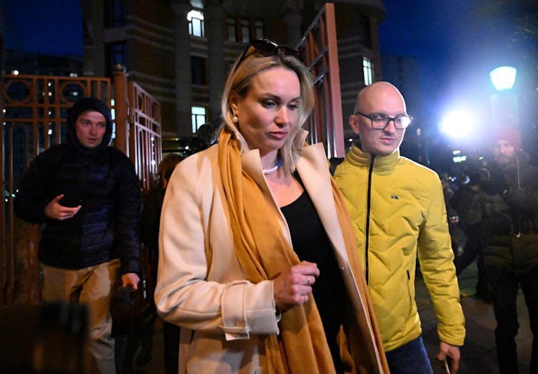 A white woman in a beige coat walks on a dark night next to a man in a yellow coat.