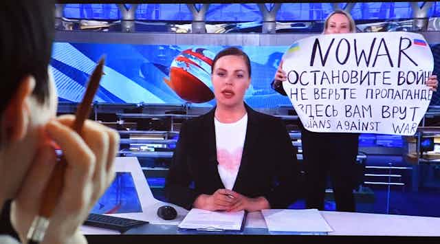 A computer screen shows a news broadcaster seated. Behind her a woman holds up a sign that says No war 