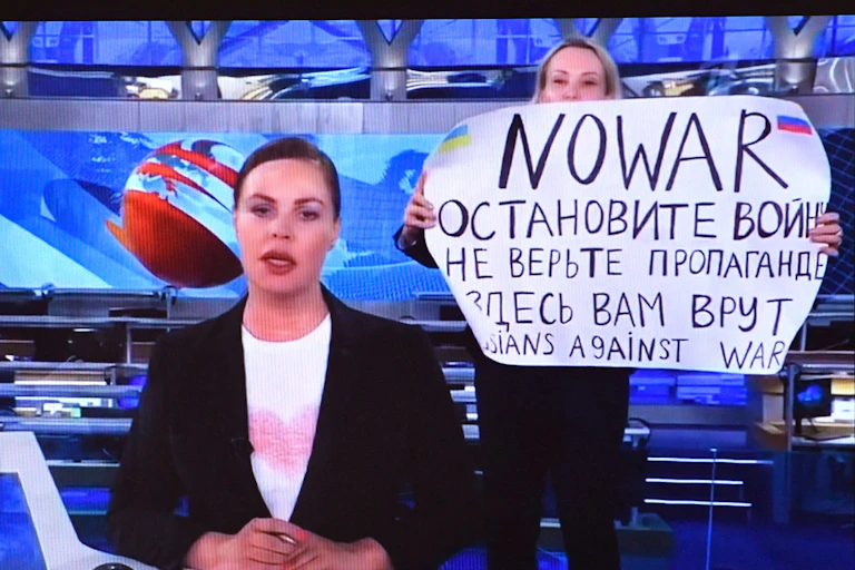 A computer screen shows a news broadcaster seated. Behind her a woman holds up a sign that says No war