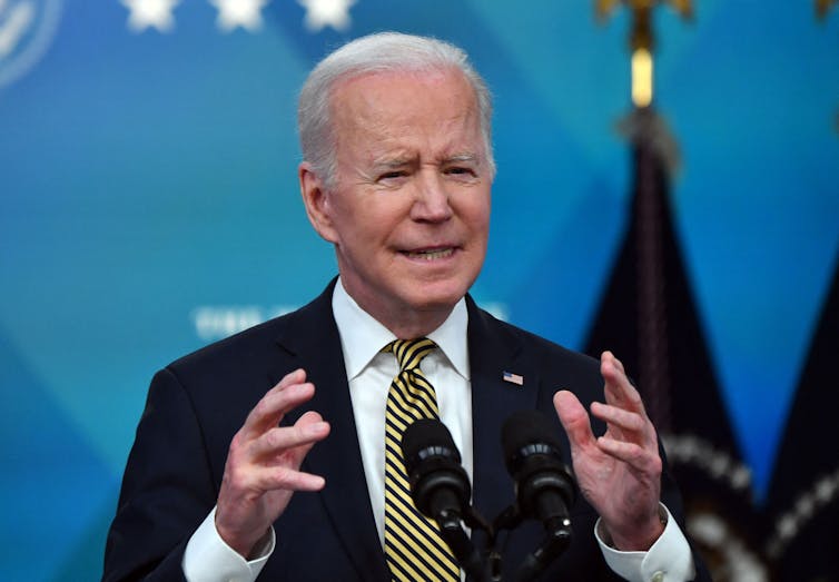 President Biden, a man with white hair and wearing a dark suit, gesturing with his hands as he gives a speech.