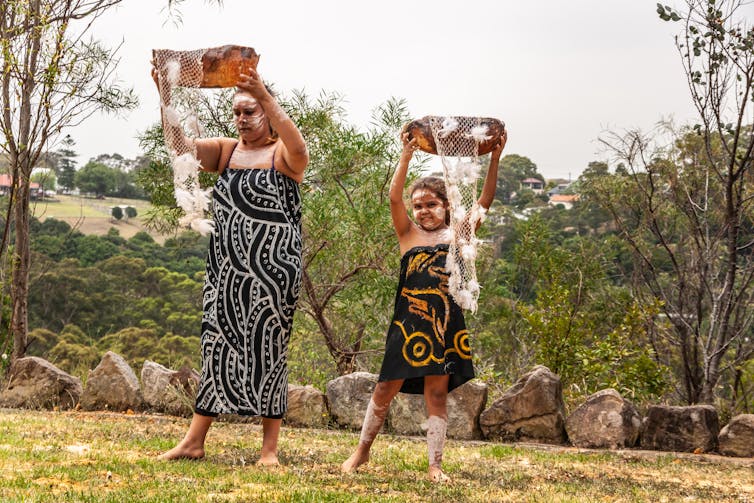 An Aboriginal person and a young child each hold a coolamon above their heads in ceremony.