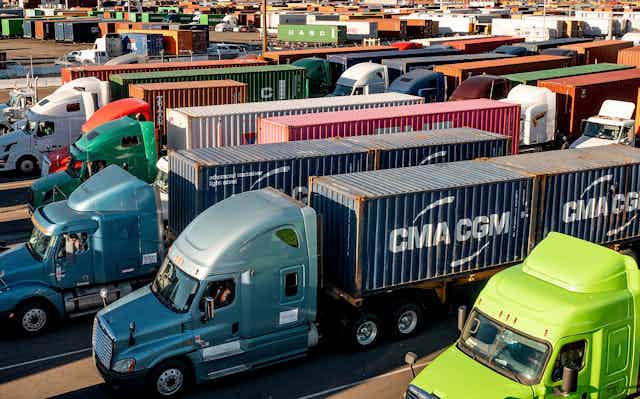 Dozens of trucks sit a traffic jam at a port with shipping containers and ships in the background.