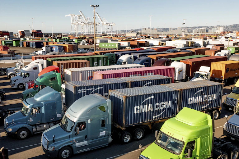 Dozens of trucks sit a traffic jam at a port with shipping containers and ships in the background.