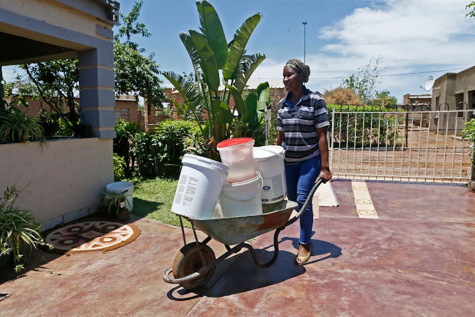 A woman pushes a wheelbarrow with buckets filled with water.