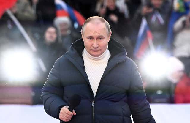 Russian president Vladimir Putin on state at a rally wearing a roll-neck sweater and anorak.