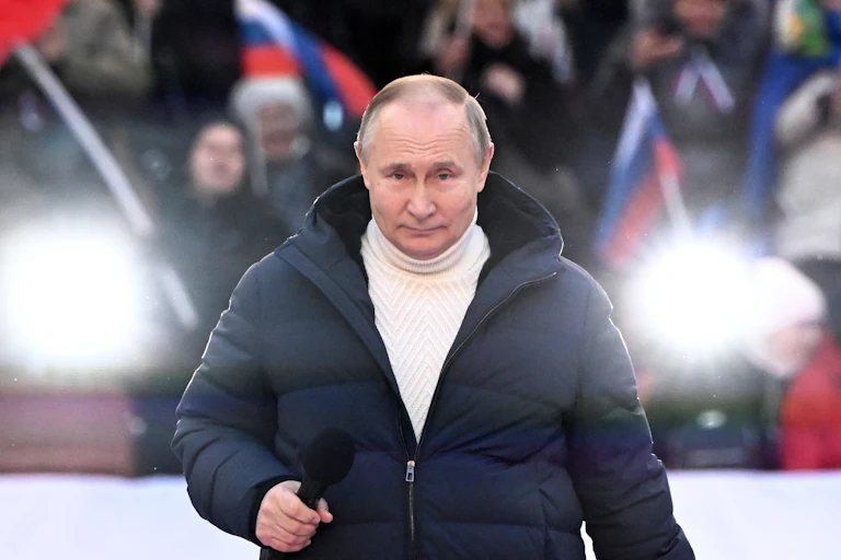 Russian president Vladimir Putin on state at a rally wearing a roll-neck sweater and anorak.