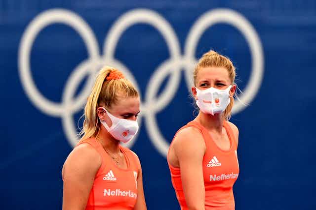 Deux athlètes passent devant les anneaux olympiques, le visage couvert d'un masque.