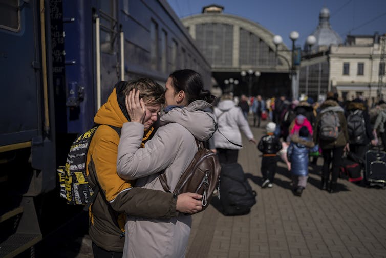 A mother on a train platform embraces her weeping son who is wearing a yellow jacket.
