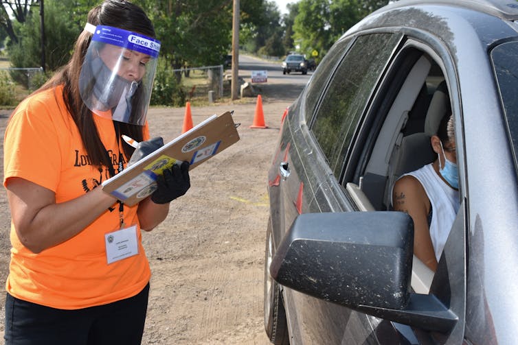 A person wearing a mask and face shield writes on a clipboard while speaking with a person in a car