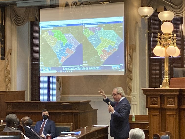 A man gestures at a screen showing two maps of political districts in South Carolina