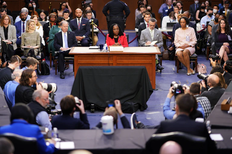 Ketanji Brown Jackson sits at a wooden table by herself, surrounded by people seated on both sides and a row of photographers in front of her