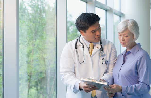A doctor wearing a white coat, with a stethoscope around his neck talking to an elderly woman.