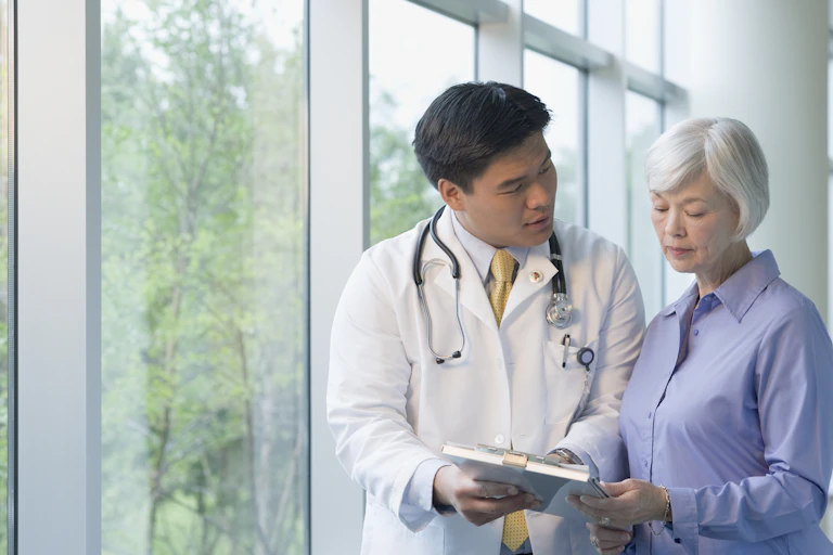 A doctor wearing a white coat, with a stethoscope around his neck talking to an elderly woman.