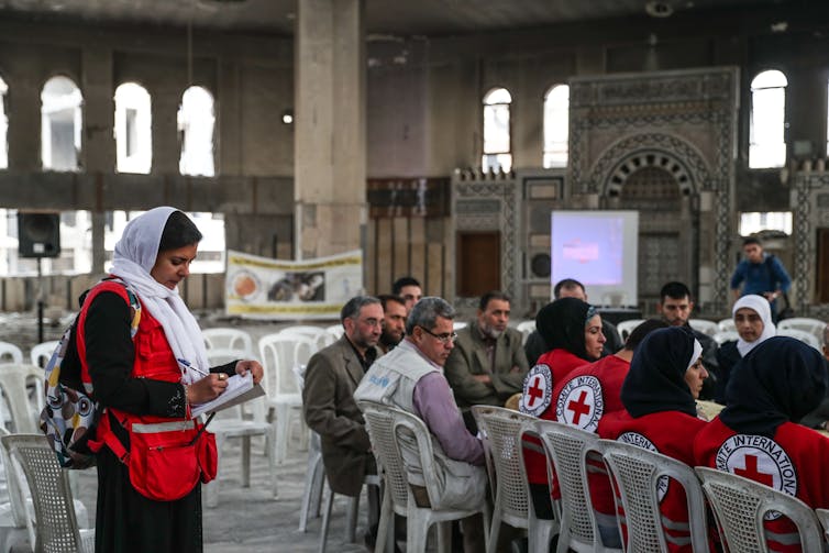 A woman in a hijab and a red vest writes on a clipboard, while other aid workers are seated at a long table beside her in a mosque