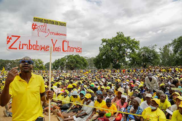 A holds a placard with the words 'Zimbabwe backs Chamisa' in front of thousanfs of people sitting on the ground at an opposition election campaign rally. 