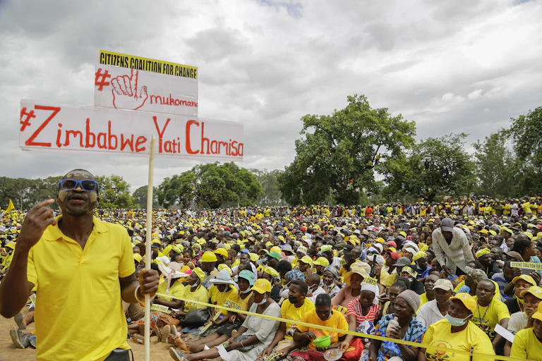 A holds a placard with the words 'Zimbabwe backs Chamisa' in front of thousanfs of people sitting on the ground at an opposition election campaign rally.