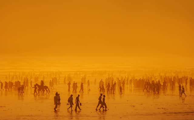 Los of people on a beach in Pakistan at sunset, some riding horses. The air is hazy, so people turn into lighter and lighter silhouettes closer to the ocean behind them.