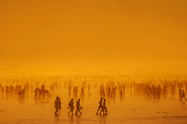 Los of people on a beach in Pakistan at sunset, some riding horses. The air is hazy, so people turn into lighter and lighter silhouettes closer to the ocean behind them.