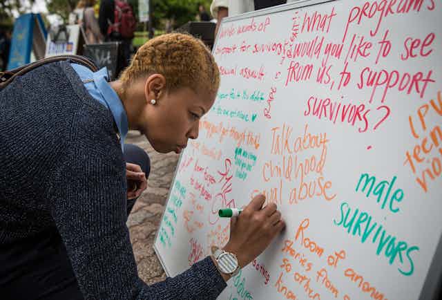 A Black woman writes on a whiteboard filled with sentiments on sexual assault. 