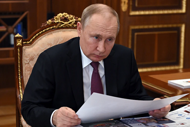 A balding man holds documents in his hands and looks over them as he sits in an ornate chair.