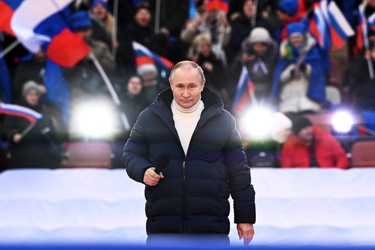 A balding man in a white turtleneck and dark parka holds a microphone as a crowd waves flags behind him.